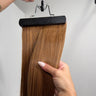 A straight hair extension piece in a warm medium brown shade, held on a black hanger by a hand with manicured nails, against a plain light gray background.