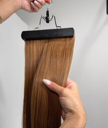 A straight hair extension piece in a warm medium brown shade, held on a black hanger by a hand with manicured nails, against a plain light gray background.
